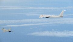 A military jet flies in close proximity to a larger reconnaissance aircraft over a cloud-filled sky.