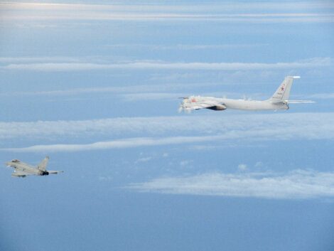 A military jet flies in close proximity to a larger reconnaissance aircraft over a cloud-filled sky.