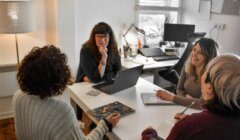 Four people sitting around a desk in an office, engaged in a meeting. There are laptops, notebooks, and a desk lamp in view.