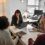 Four people sitting around a desk in an office, engaged in a meeting. There are laptops, notebooks, and a desk lamp in view.