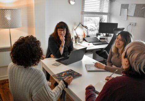 Four people sitting around a desk in an office, engaged in a meeting. There are laptops, notebooks, and a desk lamp in view.