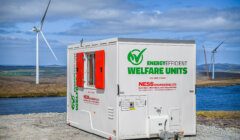 A white energy-efficient welfare unit from Ness Engineering Ltd on a gravel surface with wind turbines and a body of water in the background under a partly cloudy sky.