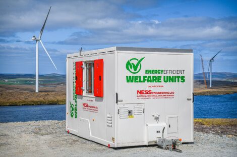 A white energy-efficient welfare unit from Ness Engineering Ltd on a gravel surface with wind turbines and a body of water in the background under a partly cloudy sky.