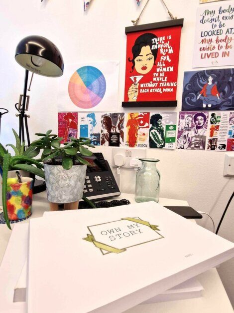 A brightly decorated workspace featuring a "Own My Story" notebook on the desk, a table lamp, potted plants, colorful drink, and art posters on the wall, including a color wheel and inspiring quote.