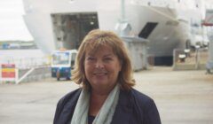 A woman with shoulder-length hair is standing in front of a large ship in a port area. She is smiling and wearing a dark jacket and light-colored scarf.