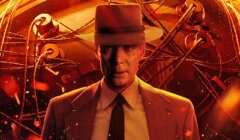 A man in a suit and hat stands in front of a large mechanical device, illuminated by fiery, orange light with wires and components visible.