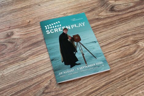 A magazine titled "Screen Play" rests on a wooden surface. The cover shows a person in a dark cloak using an old-fashioned camera. Event dates listed: 29 August - 3 September 2023.