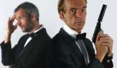 Two men in formal wear: one in focus holding a handgun, looking at the camera; the other out of focus, standing with a hand to his forehead.