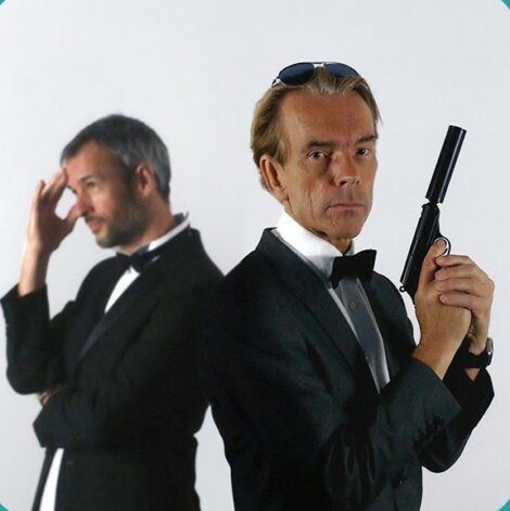 Two men in formal wear: one in focus holding a handgun, looking at the camera; the other out of focus, standing with a hand to his forehead.