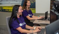 Two women wearing headsets work at desks with computers. They are both smiling and wearing purple shirts with an unreadable white logo. A scenic landscape image is in the background.