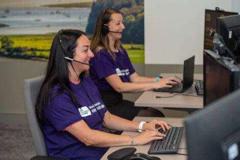 Two women wearing headsets work at desks with computers. They are both smiling and wearing purple shirts with an unreadable white logo. A scenic landscape image is in the background.