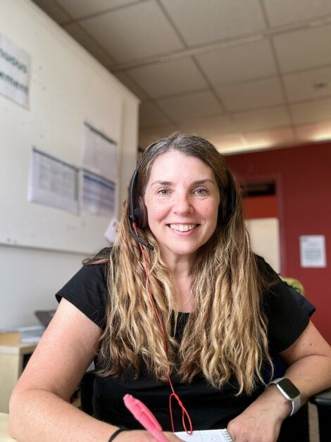 A woman with long hair, wearing a black shirt and a headset, smiles while sitting at a desk with a pen in hand. She appears to be in an office environment.