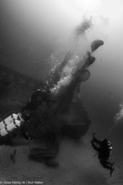 Two scuba divers inspect a submerged shipwreck underwater, with bubbles rising and gear attached.