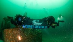 Two scuba divers hold a "Ghost Fishing UK" banner underwater, near a submerged structure, with two other divers in the background.