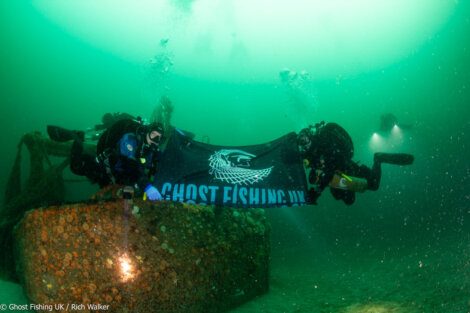 Two scuba divers hold a "Ghost Fishing UK" banner underwater, near a submerged structure, with two other divers in the background.