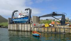 A blue boat is being lifted by a hoist on a dock. A smaller boat floats nearby in the water. Industrial equipment and buildings are visible in the background.