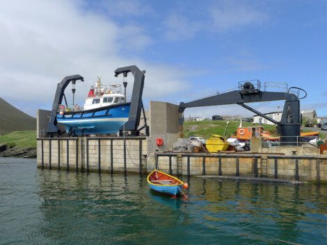 A blue boat is being lifted by a hoist on a dock. A smaller boat floats nearby in the water. Industrial equipment and buildings are visible in the background.