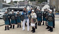 A group of people wearing green and white costumes with shields and helmets stand around a man in white clothing covered in patches, on a paved street. A white bus and buildings are visible in the background.