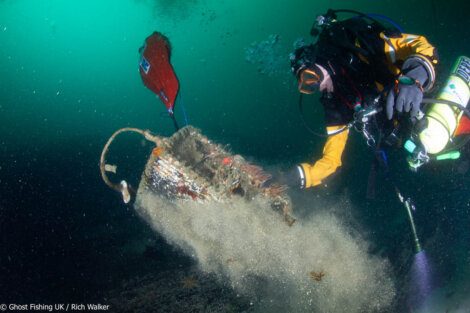 A diver in a yellow suit inspects a submerged fishing basket, releasing silty debris as it is lifted by an orange buoy underwater.