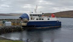 A blue and white ferry is docked at a rocky pier, with a small road leading to the vessel. The background features a cloudy sky and a hilly landscape.