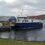 A blue and white ferry is docked at a rocky pier, with a small road leading to the vessel. The background features a cloudy sky and a hilly landscape.