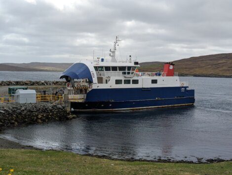 A blue and white ferry is docked at a rocky pier, with a small road leading to the vessel. The background features a cloudy sky and a hilly landscape.