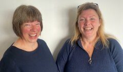 Two women smiling and standing side by side in front of a plain white background. Both are wearing dark-colored tops. One has short brown hair, and the other has longer blonde hair.