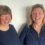 Two women smiling and standing side by side in front of a plain white background. Both are wearing dark-colored tops. One has short brown hair, and the other has longer blonde hair.