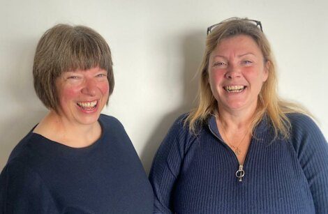 Two women smiling and standing side by side in front of a plain white background. Both are wearing dark-colored tops. One has short brown hair, and the other has longer blonde hair.