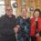 Three people are standing together indoors, all wearing event badges. The person on the left is wearing glasses and a black jacket, the person in the middle is in a dark patterned outfit, and the person on the right is in a red and black shawl.