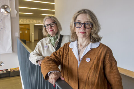 Two women with glasses stand on a balcony indoors, leaning against a railing, with the word "SCREENPLAY" partially visible in the background.