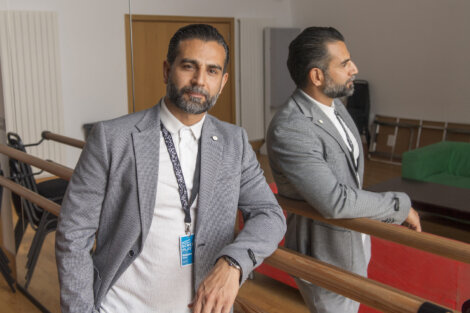 A man in business attire stands beside a mirrored wall, wearing a lanyard with a name tag. He is in a room with wooden flooring and some furniture in the background.