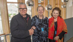 Three people are standing together indoors, all wearing event badges. The person on the left is wearing glasses and a black jacket, the person in the middle is in a dark patterned outfit, and the person on the right is in a red and black shawl.