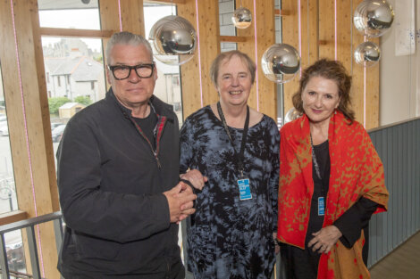 Three people are standing together indoors, all wearing event badges. The person on the left is wearing glasses and a black jacket, the person in the middle is in a dark patterned outfit, and the person on the right is in a red and black shawl.