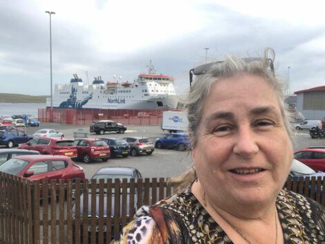 A woman with gray hair smiles at the camera with a NorthLink ferry and several parked cars in the background.