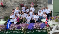 A group of people wearing matching t-shirts pose for a photo outdoors in front of a stone wall. They are surrounded by colorful flowers and flags, and are seated on and around picnic tables.