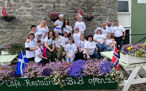 A group of people wearing matching t-shirts pose for a photo outdoors in front of a stone wall. They are surrounded by colorful flowers and flags, and are seated on and around picnic tables.