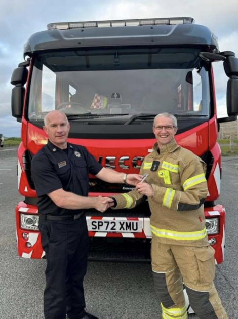 Two firefighters stand in front of a fire truck. One wears a black uniform and the other a full firefighting suit. They are shaking hands and smiling.