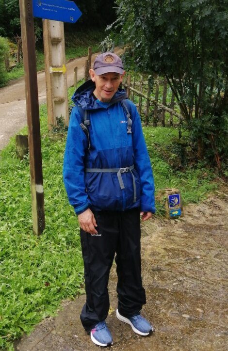 A person dressed in a blue rain jacket, black pants, and a cap stands on a wet path surrounded by greenery with a wooden post and sign in the background.