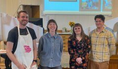 Four people stand indoors in front of a wooden desk and wall-mounted screen displaying framed pictures. One person wears an apron, and two hold hands. They smile at the camera.