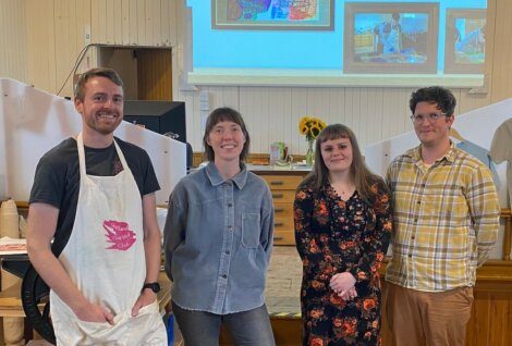 Four people stand indoors in front of a wooden desk and wall-mounted screen displaying framed pictures. One person wears an apron, and two hold hands. They smile at the camera.