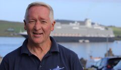 An older man stands outdoors smiling, wearing a "NorthLink Ferries" shirt. A large cruise ship is visible in the background.