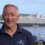 An older man stands outdoors smiling, wearing a "NorthLink Ferries" shirt. A large cruise ship is visible in the background.