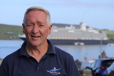 An older man stands outdoors smiling, wearing a "NorthLink Ferries" shirt. A large cruise ship is visible in the background.