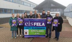 A group of people stand outside a building holding banners and signs advocating for fair pay and job protection, representing the Educational Institute of Scotland, Shetland, and UHI.