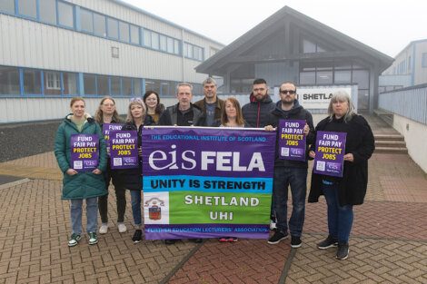 A group of people stand outside a building holding banners and signs advocating for fair pay and job protection, representing the Educational Institute of Scotland, Shetland, and UHI.