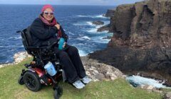 A person in a power wheelchair is seated on a grassy cliff edge overlooking a rocky coastline and the ocean. The person is wearing sunglasses and a pink bandana.