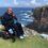 A person in a power wheelchair is seated on a grassy cliff edge overlooking a rocky coastline and the ocean. The person is wearing sunglasses and a pink bandana.