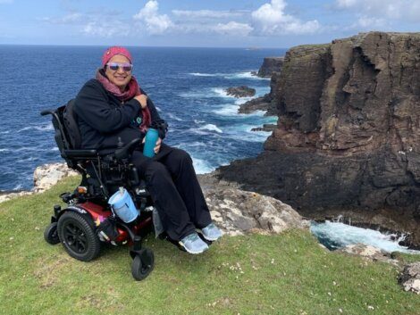 A person in a power wheelchair is seated on a grassy cliff edge overlooking a rocky coastline and the ocean. The person is wearing sunglasses and a pink bandana.