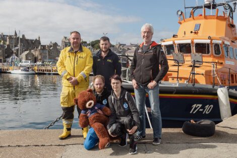 Five individuals pose on a dock in front of an orange and blue boat. One person holds a large teddy bear. The background features a waterfront with buildings and more boats.